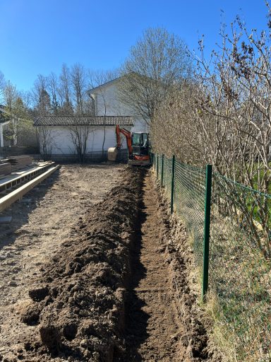 Grävd ränna längs en tomt i solljus, med ett hus och staket i bakgrunden.